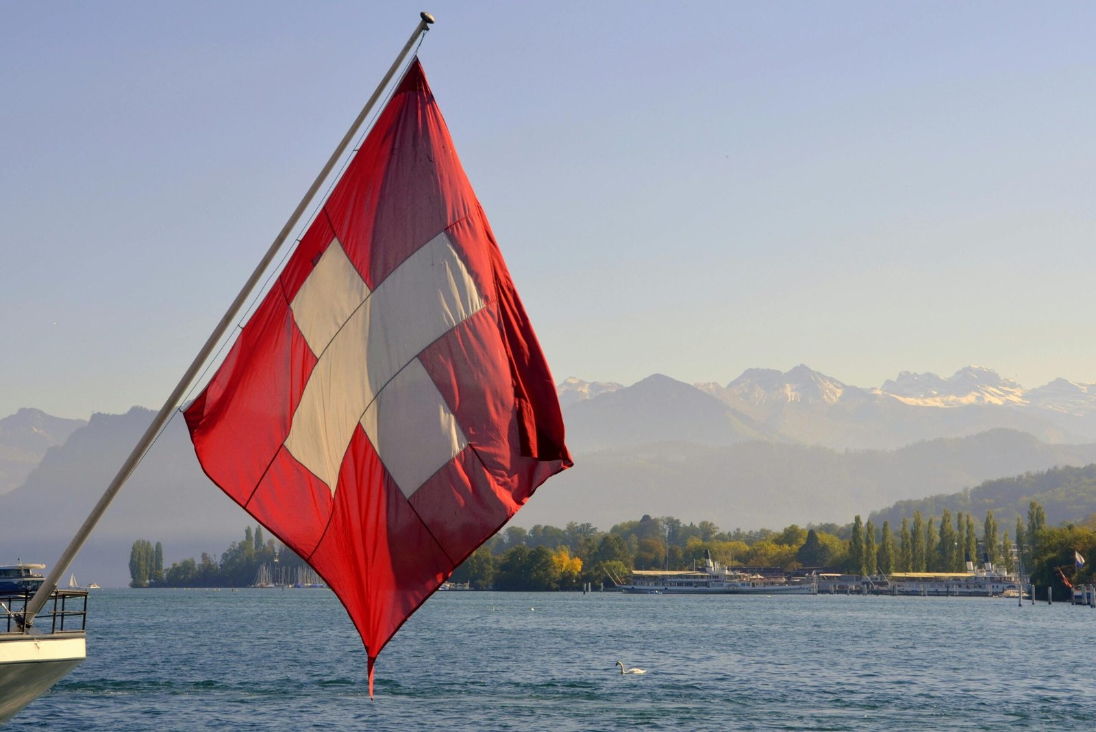 Swiss flag on a flagpole near a serene lake with breathtaking mountain backdrop in Switzerland.