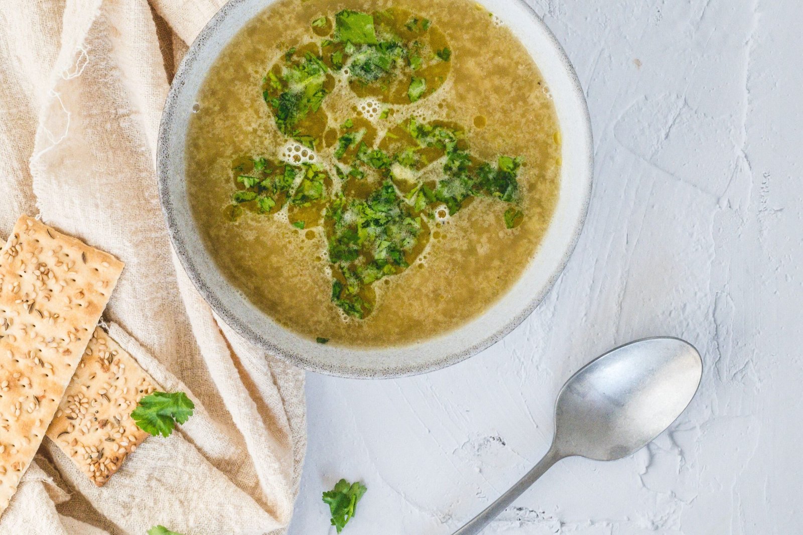 A top-down view of a bowl of green herb soup with crackers, set on a beige linen cloth.