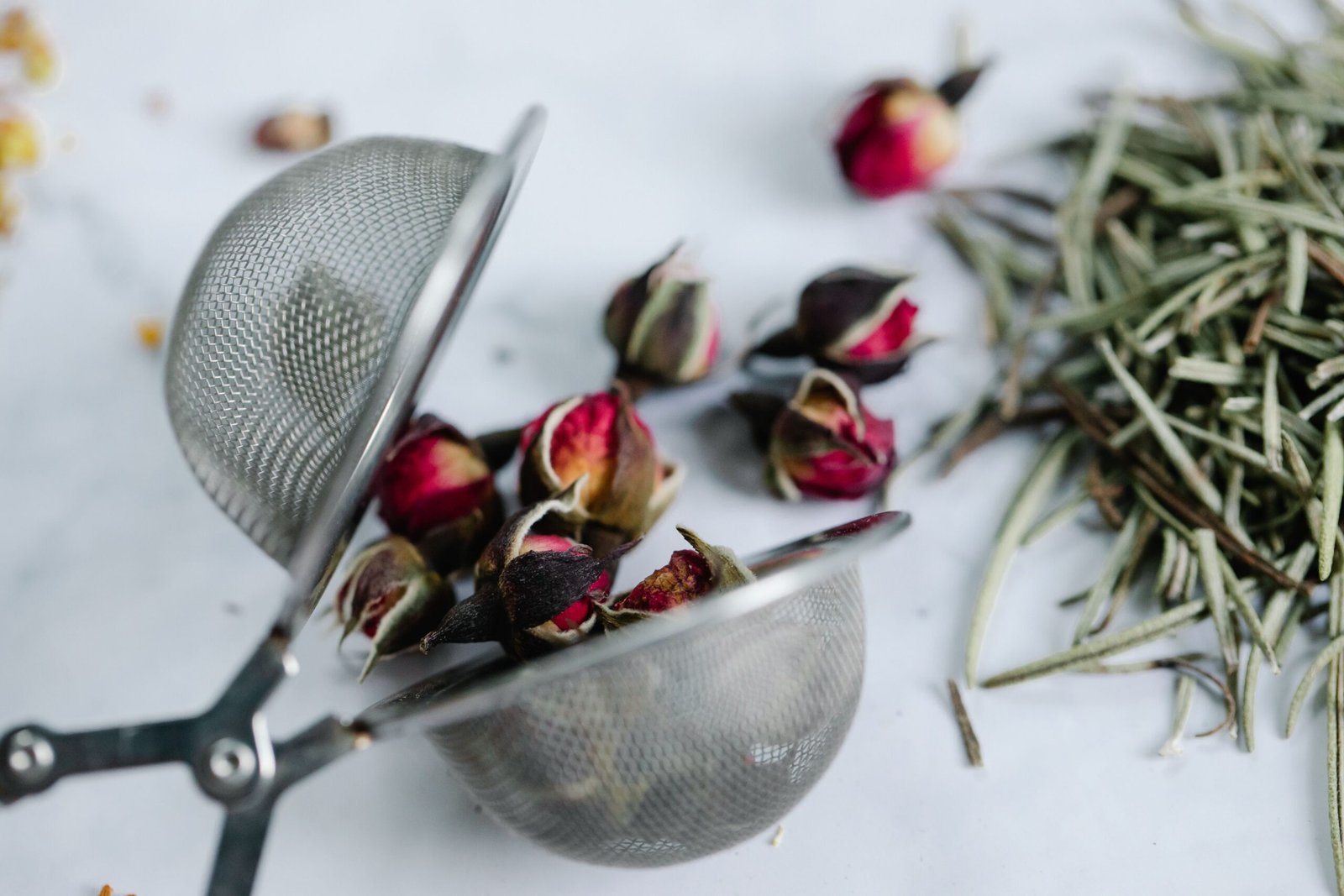 Close-up of rosebuds and herbs in a stainless steel tea strainer on a white surface.