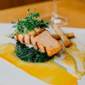 Appetizing grilled salmon steak with green spinach and french fries with sauce served on white plate on table with wine glass