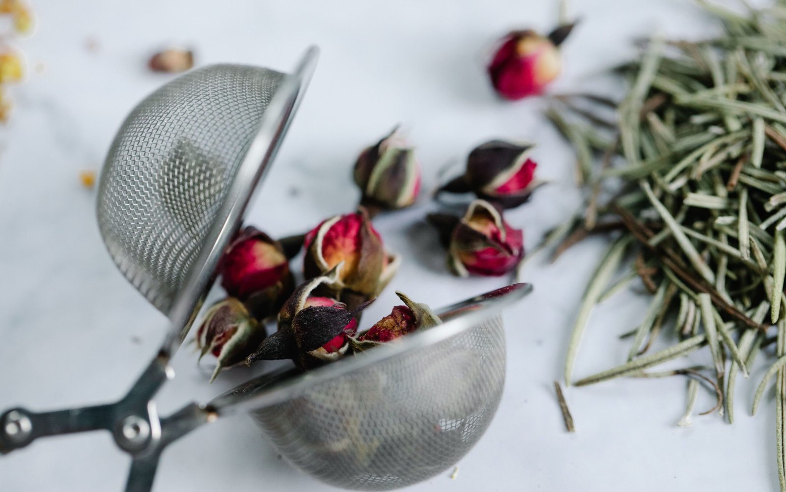 Close-up of rosebuds and herbs in a stainless steel tea strainer on a white surface.